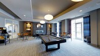 This single-floor amenity lounge is thoughtfully designed to offer a dynamic and social environment for recreation and relaxation. At the heart of the space sits a full-sized billiards table with a sleek dark finish, positioned beneath large, globe-shaped pendant lights that provide focused illumination while contributing to the room's contemporary aesthetic.

The space features a mix of distinct zones. A lounge area toward the back wall includes a mounted flat-screen TV framed by a textured stone accent wall and built-in shelving with warm wooden tones, creating a comfortable retreat for casual viewing. A foosball table adds an additional recreational element nearby. High-top tables and bar-style chairs are arranged along the perimeter, offering informal seating throughout.

Natural light enters through floor-to-ceiling windows and glass doors dressed with dark curtain panels, connecting the interior to an outdoor area and lending the room an open, airy quality. The patterned carpet, recessed lighting, and tray ceiling details add depth and a polished finish. Together, these elements create a well-appointed lounge that balances energetic recreation with relaxed social interaction.
