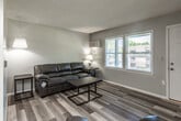This single-floor unit is designed with a functional, open layout that connects the main living areas seamlessly. The living room serves as the central gathering space, featuring light wood-look laminate flooring, neutral gray walls, and a textured popcorn ceiling. A wall-mounted HVAC unit provides climate control, while a window with horizontal blinds allows natural light to filter in and offers views of the surrounding exterior. The space is anchored by a dark leather sofa and complementary side and coffee tables.

The kitchen is equipped with rich mahogany-toned cabinetry, white appliances including a coil-top electric range and full-size refrigerator, and a countertop with an integrated sink. The kitchen opens directly into a dining area with access to natural light from an adjacent window. The flooring continues consistently throughout these connected spaces.

The bedroom is modestly sized, featuring a built-in closet rod and shelf for storage, light gray walls, and the same wood-look flooring. The bathroom includes a white vanity with cabinet storage, a medicine cabinet with mirror, a window for natural light and ventilation, and light gray wall tones that maintain a cohesive aesthetic throughout the unit.