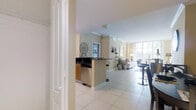 This single-floor space is designed with an open-plan layout that seamlessly connects the kitchen, dining, and living areas, creating a cohesive and functional environment. The interior is finished with large, light-colored ceramic floor tiles throughout, contributing to a clean and airy aesthetic.

The kitchen features a peninsula-style counter with dark countertops and warm wood-toned cabinetry, providing a practical workspace and casual seating opportunity. Stainless steel appliances add a modern touch, and the open design allows for easy interaction between the kitchen and surrounding living spaces.

The living area is generously proportioned and benefits significantly from large floor-to-ceiling windows that flood the space with natural light while offering expansive outdoor views. This glazing serves as a prominent architectural feature, enhancing the sense of openness throughout.

Walls are finished in a warm neutral tone, complementing the bright white ceilings adorned with crown molding details. Decorative wall art and carefully placed accessories add personality to the space. Overall, the layout and finishes combine to create a bright, welcoming, and well-connected living environment.