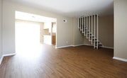 This two-floor space offers a clean, functional layout designed for comfortable everyday living. The main level is anchored by warm-toned hardwood-style flooring that runs throughout, lending a sense of continuity and warmth to the open living area. Neutral beige walls provide a versatile backdrop, complementing the light-colored baseboards and trim that frame the room with a crisp, finished look.

A prominent white open-riser staircase sits along one wall, serving as both a functional connection between floors and a subtle architectural detail. Its clean lines and white finish contrast pleasantly against the warm flooring below. A large window on the main level allows natural light to flow generously into the space, illuminating the room and offering a glimpse of outdoor greenery beyond.

The layout of the main level feels open and unobstructed, providing flexibility for a variety of furniture arrangements. A structural beam detail is visible near the ceiling, adding a layer of architectural character. Overall, the space presents a well-maintained, neutral palette that is move-in ready and easily adaptable to a range of personal styles.