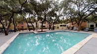 This outdoor pool area offers a well-appointed retreat designed for both relaxation and recreation. The centerpiece is a generously sized swimming pool with clear, turquoise water, framed by a classic stone coping edge and an expansive wooden sundeck lined with lounge chairs. Mature oak trees provide natural shade throughout the space, creating a park-like atmosphere that softens the surrounding architecture.

Adjacent to the pool, a covered pavilion serves as a refined gathering area, featuring natural stone columns, a warm wood-paneled ceiling, ceiling fans, and large-format stone tile flooring in muted earth tones. The pavilion's open design allows for ample airflow while offering shelter from direct sunlight. Seating areas within and around the pavilion, including woven metal chairs, provide comfortable spots to unwind.

A separate spa or hot tub is also visible near the pool, adding another dimension to the outdoor experience. The broader pool deck is equipped with additional lounge seating, and the entire area is enclosed by the surrounding buildings, creating a sense of privacy. The overall design balances modern architectural elements—clean lines, contemporary finishes, and glass accents—with natural textures, resulting in an inviting and functional outdoor amenity space.
