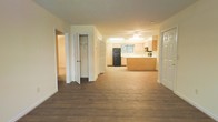 This single-floor residence offers a clean, open layout designed for comfortable everyday living. The main living area is anchored by rich, dark hardwood-style flooring that flows throughout the space, creating a warm and cohesive atmosphere. A ceiling fan overhead provides both practical airflow and a subtle design accent.

The kitchen is positioned at the heart of the home and features light wood cabinetry, a black refrigerator, and a peninsula-style breakfast bar that provides additional counter space and a natural dividing point between the kitchen and living area. A window above the sink allows natural light to filter into the cooking space, brightening the room.

A hallway connects the living area to additional rooms, lined with multiple doorways and generous storage solutions, including a dedicated closet with bifold doors — ideal for linen, utility, or general storage needs. The neutral, light-toned walls throughout the space lend a bright and airy feel that complements the darker flooring.

The overall layout is practical and well-proportioned, offering an efficient flow between the main living spaces. The combination of functional storage, open-plan design, and natural light makes this a versatile and inviting single-level home.