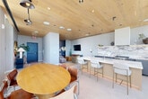 Located at 1 Florida Ave NE in Washington, DC, this sophisticated single-floor amenity space offers a seamlessly integrated open-plan layout that combines a full kitchen, dining area, and lounge. The design is anchored by a striking natural wood ceiling that adds warmth and texture, complementing the light-toned large-format tile flooring throughout. Floor-to-ceiling windows wrap around the space, flooding the interior with abundant natural light and framing sweeping panoramic views of the Washington, DC skyline and surrounding greenery — a truly standout feature of this elevated space.

The fully equipped kitchen is a centerpiece of refined craftsmanship, showcasing an expansive island and backsplash finished in dramatic book-matched marble with bold veining in warm whites, grays, and gold tones. Matte slate-blue lower cabinetry is paired with clean white upper cabinets, and a professional-grade range anchors the cooking area. Bar-height seating along the island creates an inviting space for gathering.

Adjacent to the kitchen, a round wood dining table with leather and steel chairs provides a casual yet elegant setting. The lounge area features comfortable seating and a wall-mounted television. Recessed and track lighting throughout ensures versatile illumination, while direct access to an outdoor terrace with a visible pool enhances the space's amenity appeal.