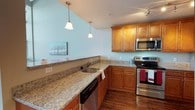 This two-story residence offers two bedrooms and two bathrooms within a well-proportioned floor plan. The kitchen stands out as a functional and attractive centerpiece, featuring warm honey-toned wood cabinetry that provides generous storage. Granite countertops with subtle veining extend across the workspace, offering ample room for meal preparation. Stainless steel appliances, including a range, microwave, and dishwasher, complement the kitchen's modern aesthetic. A double-basin undermount sink is positioned along the peninsula, which is illuminated by two pendant lights that add a refined touch. The rich hardwood flooring grounds the space, creating a warm contrast against the light blue-painted walls that give the kitchen an airy, open feel. A track lighting fixture provides additional illumination. The peninsula-style layout allows for a natural flow between the kitchen and adjacent living areas. Spread across two floors, the overall layout provides a practical separation of living and private spaces, making this residence well-suited for comfortable everyday living.