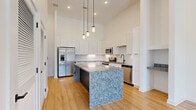 This single-floor residence is designed with a contemporary aesthetic, highlighted by soaring ceilings that amplify the sense of openness throughout the space. The kitchen serves as the centerpiece, showcasing a large, eye-catching island clad in blue-grey granite with a dark base, complemented by matching granite countertops along the perimeter. Crisp white shaker-style cabinetry extends to ceiling height, paired with a classic white subway tile backsplash and a full suite of stainless steel appliances, including a French door refrigerator, range, built-in microwave, and dishwasher. A trio of cylindrical pendant lights hangs above the island, adding refined visual interest. A built-in nook with a granite ledge is tucked beside the kitchen, offering a functional accent detail. Living areas feature light hardwood flooring, with a set of double doors drawing in natural light and outdoor views. Carpeted bedrooms offer a softer feel, each benefiting from tall ceilings and generous windows. A bathroom area features additional granite countertops, white cabinetry, open shelving, and direct access to a carpeted bedroom. Throughout, the neutral white palette and quality finishes create a clean, modern ambiance.