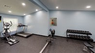 This dedicated fitness room is thoughtfully designed to support a full range of workout routines. The space is finished with calming light blue walls, dark rubber flooring ideal for high-impact activity, and recessed lighting that ensures consistent, even illumination throughout. Multiple windows fitted with white blinds allow natural light to filter in, contributing to an open and energizing atmosphere. The layout is open and spacious, providing ample room to move freely between equipment. A cable machine and weight rack support strength training, while a treadmill and rowing machine cater to cardiovascular workouts. A dumbbell rack stocked with a full range of free weights, a flat workout bench, and a tiered stability ball stand add further versatility. A wall-mounted mat and a flat-screen television are also present, enhancing both functionality and comfort. An emergency exit door is visible, reflecting practical safety considerations. The combination of varied equipment, durable finishes, and a clean, well-lit environment makes this fitness room a comprehensive and inviting space for exercise.
