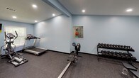 This dedicated fitness room is thoughtfully designed to support a full range of workout routines. The space is finished with calming light blue walls, dark rubber flooring ideal for high-impact activity, and recessed lighting that ensures consistent, even illumination throughout. Multiple windows fitted with white blinds allow natural light to filter in, contributing to an open and energizing atmosphere. The layout is open and spacious, providing ample room to move freely between equipment. A cable machine and weight rack support strength training, while a treadmill and rowing machine cater to cardiovascular workouts. A dumbbell rack stocked with a full range of free weights, a flat workout bench, and a tiered stability ball stand add further versatility. A wall-mounted mat and a flat-screen television are also present, enhancing both functionality and comfort. An emergency exit door is visible, reflecting practical safety considerations. The combination of varied equipment, durable finishes, and a clean, well-lit environment makes this fitness room a comprehensive and inviting space for exercise.