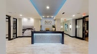 The lobby of 500 Ocean makes a strong first impression with its contemporary design and coastal aesthetic. At its centerpiece sits a bold reception desk finished with an eye-catching backlit mosaic panel that casts a cool, silvery glow across the light stone tile flooring. Above, a recessed ceiling tray is washed in vivid blue ambient lighting, complementing the crisp white surroundings and reinforcing the oceanic theme throughout the space.

The walls are tastefully appointed with carefully curated décor, including a large-scale black-and-white photographic mural depicting a serene coastal boardwalk scene, adding depth and a sense of place to the interior. Subtle sculptural accents and a potted plant atop the reception desk introduce natural elements that soften the modern lines of the space.

Recessed ceiling lighting provides clean, even illumination throughout, while a connecting corridor lined with framed artwork and wall sconces extends the polished atmosphere beyond the main entrance. The overall ambiance is professional yet welcoming, blending upscale design with a relaxed, beachside sensibility that reflects the identity of the 500 Ocean brand.