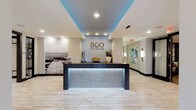 The lobby of 500 Ocean makes a strong first impression with its contemporary design and coastal aesthetic. At its centerpiece sits a bold reception desk finished with an eye-catching backlit mosaic panel that casts a cool, silvery glow across the light stone tile flooring. Above, a recessed ceiling tray is washed in vivid blue ambient lighting, complementing the crisp white surroundings and reinforcing the oceanic theme throughout the space.

The walls are tastefully appointed with carefully curated décor, including a large-scale black-and-white photographic mural depicting a serene coastal boardwalk scene, adding depth and a sense of place to the interior. Subtle sculptural accents and a potted plant atop the reception desk introduce natural elements that soften the modern lines of the space.

Recessed ceiling lighting provides clean, even illumination throughout, while a connecting corridor lined with framed artwork and wall sconces extends the polished atmosphere beyond the main entrance. The overall ambiance is professional yet welcoming, blending upscale design with a relaxed, beachside sensibility that reflects the identity of the 500 Ocean brand.