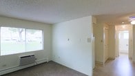 This single-floor space features a straightforward, functional layout with clean white walls and a textured popcorn ceiling. The main living area is finished with wall-to-wall carpeting, providing a comfortable underfoot feel, while the adjoining hallway transitions to a light-toned hard surface floor. A window along one wall allows natural light to filter into the room, offering a view of a grassy outdoor area. Beneath the window sits a wall-mounted air conditioning unit, ensuring climate control. The white walls are largely open, accented by a few wall outlets and a thermostat panel, keeping the space neutral and adaptable to various furnishing arrangements. A partial wall divides the living area from what appears to be a corridor leading further into the unit, hinting at additional rooms beyond. The overall ambiance is simple, bright, and move-in ready, offering a blank canvas well-suited for personalization.