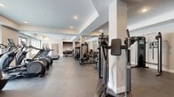 Located at 2274 Princess St in Kingston, Ontario, this well-appointed fitness facility offers an expansive, single-floor layout designed to accommodate a full spectrum of workout needs. The space is thoughtfully organized into distinct functional zones, providing ample room for both cardio training and strength work without feeling crowded.

The cardio area is lined with large windows that invite natural light and provide views of the surrounding building exterior, creating an open and energizing atmosphere. Multiple treadmills, elliptical trainers, and stair climbers are neatly arranged along the perimeter, allowing users to exercise in a bright, airy environment.

The strength training section is equally well-equipped, featuring a power rack with a flat bench, a cable crossover machine, a multi-station functional trainer, and an assortment of selectorized weight machines. A dedicated free weights area includes medicine balls, stability balls, and adjustable benches, supported by wall-mounted mirrors that enhance the sense of space and assist with form.

Underfoot, durable rubber flooring covers the high-impact training zones, while smooth concrete-style flooring extends through the cardio and machine areas. Structural columns are integrated cleanly into the layout, and recessed ceiling lighting ensures consistent, even illumination throughout. The overall design is modern and clean, with a neutral colour palette of white, grey, and beige that reinforces a professional fitness environment.