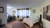 This single-floor space centers around a generously sized bedroom that combines comfort and style. The room is anchored by a large bed dressed in crisp white linens, complemented by an upholstered settee with decorative pillows positioned beneath a bank of wide windows. The windows are dressed with layered curtains — sheer white panels beneath dark ombre drapes with grommet detailing — allowing for flexible light control while bringing in abundant natural light when open.

The flooring is a distinctive herringbone parquet wood pattern, adding warmth and a classic architectural character to the space. A large area rug defines the central zone, enhancing the room's sense of coziness. A compact seating area near the window features a tufted chair and small desk or side table, offering a functional corner for reading or work.

The walls are painted a clean, neutral white, keeping the space feeling bright and open. Framed artwork is displayed on both sides of the room, adding personality and a curated aesthetic. The overall ambiance is calm and refined, with a mix of traditional and transitional design elements throughout.
