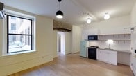 This single-floor space masterfully combines historic architectural character with contemporary design. The open-plan layout creates a seamless flow between the living area and kitchen, bathed in natural light streaming through large windows with classic black frames.

The kitchen is a standout feature, outfitted with crisp white cabinetry, a black gas range, a built-in microwave, and a charming retro-style mint green SMEG refrigerator that adds a playful pop of color. A stainless steel sink and ample counter space complete the functional yet stylish setup.

One of the most striking architectural details is the exposed brick and stone archway framing the passage between rooms, offering a nod to the building's industrial or historic origins. This raw textural element contrasts beautifully with the smooth, light-toned walls and pale hardwood floors that run throughout the space, lending warmth and continuity underfoot.

Overhead, modern pendant lighting — including a sleek matte black globe fixture and a round white flush-mount light — provides both ambient and focused illumination. Subtle ceiling beams further reinforce the building's heritage character while contributing to the elevated sense of scale. Together, these elements create an inviting, thoughtfully designed space that balances old-world charm with modern livability.