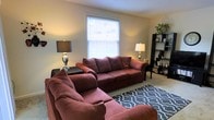 This single-story residence at Bryce De Moray Apartments offers a comfortable and well-appointed living space designed for everyday ease. The living room serves as the heart of the home, featuring neutral-toned walls that create a warm, inviting atmosphere. A centrally positioned window fitted with horizontal blinds allows natural light to filter into the room, brightening the space throughout the day while offering privacy when needed. The room comfortably accommodates generous seating, as evidenced by the spacious sofa arrangement paired with a patterned area rug that anchors the seating zone and adds visual interest to the carpeted floor. A dark shelving unit along one wall provides both display and storage opportunities, contributing to the room's organized feel. Accent lamps on either side of the seating area offer warm ambient lighting, enhancing the cozy character of the space during evening hours. The overall layout of the living area reflects a practical yet comfortable design, with sufficient room to move freely and arrange furnishings to suit personal preference. The single-floor layout ensures easy accessibility and a seamless flow throughout the residence.