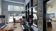This fitness center combines modern design with natural materials to create an inviting and functional workout environment. The space is distinguished by its soaring ceiling height, which contributes to an open, airy atmosphere throughout. Large floor-to-ceiling windows line the walls, flooding the interior with abundant natural light and offering unobstructed views of a neighboring outdoor pool area and mature trees beyond.

Prominent natural stone columns serve as architectural focal points, adding texture and warmth that contrasts with the sleek, contemporary finishes found elsewhere in the space. The flooring is a light-toned hardwood, complementing the overall design aesthetic while remaining practical for a fitness setting.

Cardio equipment, including a treadmill and elliptical machine, is thoughtfully arranged to take advantage of the outdoor views, enhancing the exercise experience. A wall-mounted television provides additional entertainment options. Additional strength and stretching equipment is visible toward the perimeter. Dark-framed windows and glass partitions add a refined, modern edge to the space, while the combination of stone, wood, and steel materials creates a cohesive and upscale fitness environment.