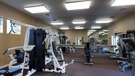 This fitness center is designed to support a comprehensive workout experience within a single-level layout. The space is outfitted with a diverse selection of strength training equipment, including multi-function cable machines and weight stations, as well as cardio machines visible toward the back of the room. The flooring features a dark, cushioned surface well-suited for exercise, providing comfort and durability underfoot.

Large, full-length mirrors line the walls, visually expanding the space and allowing users to monitor their form during workouts. The ceiling is finished with a textured surface and fitted with evenly spaced, recessed rectangular fluorescent light panels that provide consistent, bright illumination throughout the room. Ventilation is addressed with visible ceiling vents, contributing to a comfortable training environment.

The open floor plan allows for good circulation between equipment, making it accessible and functional. The warm-toned walls contrast with the darker flooring, creating a practical and focused atmosphere. Overall, the space is purposefully arranged to accommodate a range of fitness activities in a clean, well-lit setting.
