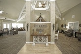 This single-story community gathering space is defined by its dramatic vaulted ceilings with exposed white beams, which lend an open, airy quality to the interior. The architectural centerpiece is a striking floor-to-ceiling brick fireplace, featuring a decorative mantel and a classic clock, anchoring the main living area with warmth and character. Adjacent to the fireplace is a white dining table with dark upholstered chairs, providing a functional space for group activities or casual dining.

The space is organized into several distinct seating zones, each thoughtfully furnished. Rich brown leather and fabric sofas, plush oversized armchairs, and patterned accent chairs are arranged throughout, offering comfortable gathering areas. A dark wood coffee table serves a central lounge configuration, while side tables with lamps provide ambient lighting.

Natural light is abundant, with numerous windows dressed in white blinds and multiple glass-paneled exterior doors that allow views of the surrounding outdoors. Earthy green walls and textured multi-toned carpet flooring unify the space. A resource area with shelving and a desk adds a practical dimension, while ceiling fans and exposed ductwork contribute to the functional, community-oriented character of the space.