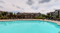 This single-floor amenity space offers residents a comprehensive wellness and recreation experience. The centerpiece of the outdoor area is a generously sized swimming pool with crystal-clear turquoise water, surrounded by a wide sundeck lined with chaise lounge chairs. A wooden pergola structure provides a shaded gathering area along the poolside, while lush landscaping and views of the surrounding residential buildings complete the outdoor ambiance.

Inside, the fitness studio is thoughtfully designed with vaulted ceilings featuring wood-paneled accents and a neutral color palette that creates an open, airy atmosphere. Large windows allow abundant natural light to fill the space while offering views of the pool and courtyard beyond. The studio is divided into functional zones: a yoga and stretching area with floor mats, a rack of colorful kettlebells, and wall-mounted mirrors for form checks, as well as a dedicated cycling zone equipped with multiple stationary spin bikes. A bold, vibrant mural adds an energetic artistic focal point to the main workout area, complemented by storage shelving for accessories and stability balls. Wall-mounted televisions and durable dark flooring throughout complete the practical, motivating environment.
