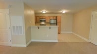 This single-floor residence offers a practical and well-appointed open-concept layout that seamlessly connects the kitchen and living areas. The kitchen is equipped with stainless steel appliances, including a refrigerator, microwave, and electric range, all complemented by warm honey-toned wood cabinetry that provides generous storage space. A wraparound breakfast bar with barstool seating extends from the kitchen, creating a natural divide between the cooking and living spaces while also serving as a convenient spot for casual dining or entertaining. The countertops are finished in a light, neutral tone that coordinates with the overall color palette of the home. Flush-mount ceiling light fixtures provide consistent overhead lighting throughout the space. The living area features soft, wall-to-wall carpeting in a warm beige tone, while the kitchen is finished with light-colored tile flooring. The walls throughout are painted in a soft, neutral beige, creating a cohesive and inviting atmosphere. Clean white baseboards add a crisp finishing detail to each room. The overall design is straightforward and functional, offering a comfortable, move-in-ready environment with a neutral palette that easily accommodates a variety of interior styles.