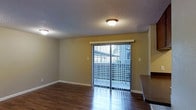 This single-floor space offers a clean and functional layout with a welcoming sense of openness. The main living area is highlighted by rich, dark hardwood-style flooring that runs throughout, providing a warm and polished foundation. A textured ceiling with flush-mount lighting fixtures provides even illumination across the room, complementing the natural light that filters in through the prominent sliding glass door fitted with vertical blinds. The sliding door serves as a key architectural feature, offering direct access to an outdoor balcony space while framing views of the surrounding exterior. The neutral-toned walls create a versatile backdrop suitable for a variety of furnishing styles. Clean white baseboards add a crisp finishing detail throughout. Adjacent to the living area, a countertop edge is visible, suggesting a connected kitchen or dining space that contributes to the open feel of the floor plan. The overall ambiance is practical yet comfortable, with a layout that encourages easy flow between the living and adjacent areas. Natural light plays an important role in the character of the space, making it feel airy and inviting throughout the day.