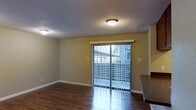 This single-floor space offers a clean and functional layout with a welcoming sense of openness. The main living area is highlighted by rich, dark hardwood-style flooring that runs throughout, providing a warm and polished foundation. A textured ceiling with flush-mount lighting fixtures provides even illumination across the room, complementing the natural light that filters in through the prominent sliding glass door fitted with vertical blinds. The sliding door serves as a key architectural feature, offering direct access to an outdoor balcony space while framing views of the surrounding exterior. The neutral-toned walls create a versatile backdrop suitable for a variety of furnishing styles. Clean white baseboards add a crisp finishing detail throughout. Adjacent to the living area, a countertop edge is visible, suggesting a connected kitchen or dining space that contributes to the open feel of the floor plan. The overall ambiance is practical yet comfortable, with a layout that encourages easy flow between the living and adjacent areas. Natural light plays an important role in the character of the space, making it feel airy and inviting throughout the day.