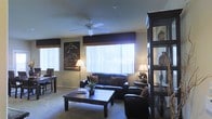 This single-floor space exudes a warm, inviting atmosphere complemented by a distinctive Western-inspired aesthetic. The living area serves as the heart of the home, thoughtfully arranged around a rich leather sofa and matching armchair that anchor the room with a sense of comfort and style. A sturdy wooden coffee table sits at the center, providing both function and visual grounding to the seating arrangement. A ceiling fan overhead ensures year-round comfort while adding a classic touch to the space. Large windows dressed with vertical blinds and dark valances flood the room with abundant natural light, creating a bright and airy feel throughout. A Western-themed art poster adds a bold focal point to the wall, reinforcing the space's character and personality. A dark, open display shelving unit along one side of the room showcases decorative accessories, adding depth and visual interest. Soft carpeting underfoot contributes to the overall warmth of the interior. A dining or workspace area with a table and chairs is visible nearby, offering a versatile zone within the open layout. The cohesive dark wood and leather palette throughout gives the space a unified, intentional design sensibility.