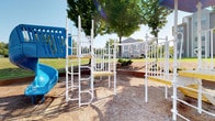 This community space combines an inviting indoor clubhouse with an outdoor playground, offering amenities designed for both leisure and productivity. The centerpiece of the interior is a spacious great room characterized by soaring vaulted ceilings adorned with prominent exposed wood beams, creating a warm, architectural focal point. Tall, multi-pane windows line the walls, flooding the room with abundant natural light and offering views of the surrounding greenery. A white-mantled fireplace anchors one end of the great room, complemented by hardwood-style flooring and wainscoting details that add a refined, classic touch throughout.

Adjacent to the great room is a kitchenette area equipped with dark cabinetry, a stainless steel refrigerator, and a light-topped island counter — well-suited for community gatherings. A dedicated business center features a shared workstation desk with multiple computer setups, ergonomic seating, and its own vaulted ceiling with exposed beams, maintaining the cohesive architectural style. Natural light enters through a glass-paned door, keeping the workspace bright and welcoming.

Outside, the playground features a multi-component white metal play structure with climbing ladders, platforms, handholds, and a blue slide, all set on a cushioned wood-chip surface beneath mature shade trees, providing an engaging outdoor recreation area.