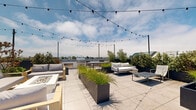 Located in the Mar Vista neighborhood of Los Angeles, this rooftop deck at 12602 Matteson Ave offers a well-appointed outdoor communal space atop a modern multi-story building. The rooftop is designed for both relaxation and entertainment, featuring multiple seating arrangements with contemporary outdoor sofas, lounge chairs, and low-profile coffee tables arranged across a large, clean-lined tiled terrace. Overhead, strands of globe string lights are suspended between posts, adding a warm, inviting ambiance for evening gatherings. Tall, dark-toned rectangular planters filled with ornamental grasses and lush shrubs are distributed throughout the space, providing natural greenery and visual separation between seating zones. An outdoor kitchen area equipped with a stainless steel grill and dining table anchors one end of the deck, making it ideal for al fresco dining. The surrounding railings are finished in a sleek dark metal, complementing the building's crisp white modern exterior. Open walkways and stairways connect the rooftop to the building's interior courtyard below. Panoramic views of the Los Angeles skyline, treetops, and surrounding neighborhoods extend in multiple directions, taking full advantage of the elevated position under the wide Southern California sky.