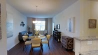 This single-floor space showcases an open-concept design that seamlessly connects the dining and living areas, creating a cohesive and functional layout. The dining area is anchored by a round table surrounded by cushioned chairs in neutral tones, illuminated by an elegant chandelier that adds a refined touch overhead. Dark hardwood flooring runs throughout, providing a rich contrast against the light, neutral-toned walls and contributing to a warm, grounded atmosphere. Natural light filters in through windows dressed with layered curtains, brightening the space and adding depth. The living area, visible from the dining space, features a comfortable sofa and accent pieces that complement the overall aesthetic. Along one wall, a dark media console serves as a functional centerpiece, flanked by decorative accessories and wall art that add personality to the room. A rustic wooden chest with metal accents introduces a charming, eclectic element. The overall ambiance is inviting and thoughtfully curated, balancing contemporary furnishings with warm, classic details that make the space feel both stylish and livable.