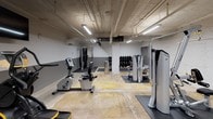 Located on N Church St in Spartanburg, South Carolina, this fitness facility occupies a single open floor with an industrial aesthetic that blends raw architectural character with modern gym amenities. The space features exposed concrete ceilings with visible conduit and ductwork, painted brick walls, and a two-tone grey and white color scheme that gives the interior a clean, utilitarian feel. Bright fluorescent and LED lighting ensures the entire floor is well-illuminated throughout.

The cardio zone is lined with a strong selection of True Fitness equipment, including treadmills, elliptical trainers, a recumbent bike, and a stair climber. Multiple wall-mounted flat-screen televisions are positioned throughout this area to keep members engaged during workouts. The strength training section offers a variety of cable and resistance machines, along with a dumbbell rack stocked with a full range of free weights. Rubber floor mats are strategically placed beneath equipment to provide safety and protection.

Additional amenities include a water dispenser and hydration station conveniently located near the entrance, as well as designated storage areas. The facility is accessed through a set of dark green glass-paneled doors and is clearly marked with safety signage, including illuminated exit indicators. The overall layout provides an efficient and functional workout environment suitable for a range of fitness levels and training styles.