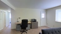 This single-floor office space offers a clean, functional environment designed to support productive work. The main workspace is anchored by a light wood-toned desk paired with a black ergonomic office chair, providing a comfortable and practical setup. A metal desk lamp and a small clock sit atop the desk, adding useful utility to the workspace. Beneath the desk, a multi-drawer metal filing cabinet offers ample document storage, while a separate gray storage cabinet positioned nearby houses additional equipment, including a multifunction printer. The room features neutral, light-colored walls that contribute to an open and airy atmosphere, complemented by wall-to-wall carpeting in a warm beige tone that softens the overall aesthetic. A white paneled door with a frosted window allows natural light to filter into the space, brightening the interior and creating a welcoming ambiance. A portion of a dark upholstered chair is visible in the foreground, suggesting an additional seating area within the room. The overall layout is efficient and well-organized, making this space well-suited for administrative, professional, or home office use.