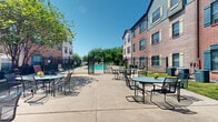 Located at 12400 Castlebridge Dr in Jersey Village, Texas, this outdoor amenity space is a well-appointed communal area designed for relaxation and social gathering. The centerpiece of the space is a generously sized resort-style swimming pool with a smooth, curved design and built-in water features, including cascading water jets that add a lively ambiance. Adjacent to the pool is a separate spa, offering a dedicated space for leisure. The pool area is enclosed by ornamental black iron fencing with a gated entry, ensuring both security and aesthetic appeal.

Surrounding the pool, a spacious concrete patio provides ample room for outdoor furniture, with multiple round tables and chairs arranged throughout for comfortable outdoor dining and lounging. The courtyard extends beyond the pool enclosure into a broader outdoor seating area, framed by the multi-story brick exterior of the buildings. The architecture features a mix of red and tan brick with teal and dark-hued shuttered windows, creating a classic and cohesive aesthetic.

Mature shade trees are scattered throughout the property, providing natural relief from the Texas sun. The open-sky setting allows abundant natural light to fill the space, contributing to its bright and welcoming atmosphere. The grounds are well-maintained, with potted plants and flower accents adding color and charm to the overall environment.