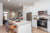 Located near the Mall of Georgia in Buford, GA, this single-floor residence offers a well-appointed, open-concept layout with contemporary finishes and a clean, modern aesthetic. The kitchen serves as a central highlight, featuring white shaker cabinetry, stainless steel appliances, granite countertops, and a peninsula island with bar seating — ideal for casual dining and entertaining. Light wood-style flooring flows through the main living and kitchen areas, complementing the bright white walls and recessed lighting overhead.

The living area is generously proportioned, with access to an outdoor space through a glass door flanked by tall windows that draw in abundant natural light. The elevated ceiling height in this area enhances the sense of openness throughout.

The bedroom is a comfortable retreat, carpeted for warmth, and features large windows with sheer drapery that allow natural light while maintaining privacy. An adjoining bathroom offers a granite-topped vanity with white cabinetry and a large mirror, continuing the cohesive design language found throughout the unit.

A dedicated laundry area houses a stacked washer and dryer alongside a closet with a hanging rod, providing practical storage and convenience. Overall, the space presents a refined, move-in-ready environment with consistent modern detailing from room to room.