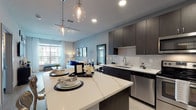 This single-floor residence is thoughtfully designed with a modern aesthetic, combining functionality and style throughout its open layout. The kitchen serves as a central highlight, featuring dark wood cabinetry, a white marble-veined quartz island with bar seating, and a full suite of stainless steel appliances including a range, microwave, dishwasher, and refrigerator. A white subway tile backsplash and glass globe pendant lights add refined detail, while light wood-style flooring flows seamlessly into the adjoining living area.

The living room is bright and welcoming, benefiting from large windows that frame outdoor views and fill the space with natural light. Neutral gray walls provide a versatile backdrop complemented by light curtains and contemporary furnishings.

The primary bedroom features a floor-to-ceiling upholstered accent wall and coordinating nightstands, creating a polished, hotel-like atmosphere. A second bedroom offers a large window with outdoor views, patterned drapery, and ample dresser space. The bathroom is finished with granite countertops, dark cabinetry, a large vanity mirror, and a glass-enclosed shower, all set on light tile flooring. The entry area includes a practical key hook station near the front door.