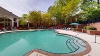 The outdoor pool area at 4800 Alexander Valley Drive in Charlotte, NC, offers a well-appointed amenity space designed for relaxation and recreation. The centerpiece is a generously sized freeform swimming pool with clear turquoise water, framed by brick coping that adds a classic, polished aesthetic. The pool features gradual entry points with stainless steel handrails, and depth markers are clearly visible, indicating a shallow entry that deepens toward the interior. An attached spa or wading area is positioned adjacent to the main pool, providing a dedicated space for leisure.

The surrounding deck is finished in a neutral concrete surface, offering ample room for circulation and lounging. Numerous blue reclining lounge chairs are arranged along the pool's perimeter, providing plentiful seating for sunbathing. Shaded areas are available via a blue market umbrella with accompanying seating, offering relief from direct sunlight. A brick retaining wall borders one side of the deck, adding architectural definition to the space.

The entire area is enveloped by mature, leafy trees that provide natural shade and privacy, creating a secluded, resort-like atmosphere. The brick clubhouse building visible in the background complements the traditional architectural character of the community.