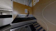 This single-level underground parking facility offers a generously sized, open-plan layout accommodating multiple vehicle spaces marked with clearly painted directional arrows and stall lines on a polished concrete floor. The structure is constructed with exposed cast-in-place concrete walls, columns, and ceilings, lending it a utilitarian yet durable character. Overhead, a network of mechanical pipes, conduits, and fluorescent strip lighting runs throughout, ensuring consistent illumination across the entire level. A staircase with metal railings provides access to upper floors, and safety features such as illuminated exit signs and a fire suppression system are visibly integrated into the ceiling infrastructure. Safety bollards in yellow mark key structural columns throughout the space.

A standout amenity within this parking level is a dedicated pet wash station, branded as the "Pet Spa" by Cascades. Set against bold golden-yellow walls adorned with the Cascades logo and circular graphic motifs, the station features a large, deep stainless steel wash tub equipped with a faucet and a coiled handheld spray hose, along with built-in steps to assist pets in accessing the tub. Posted wash policies ensure proper usage of the facility, making this a well-considered and practical addition to the overall space.