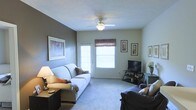 This single-floor space offers a warm and functional layout designed for comfortable everyday living. The living room serves as the central gathering area, featuring plush wall-to-wall carpeting that adds a sense of warmth underfoot. A ceiling fan with an integrated light fixture is centrally mounted, providing both air circulation and ambient lighting throughout the room. The space is brightened by a large window dressed with a decorative valance, allowing natural light to filter in, accompanied by an exterior-facing door that enhances accessibility and ventilation. The room accommodates generous seating with multiple upholstered sofas, offering ample space for relaxation and entertaining. A media console with a flat-screen television anchors one wall, creating a dedicated entertainment area. The walls feature a two-tone color scheme, with a deep accent wall providing visual contrast against the lighter surrounding walls. Framed artwork is thoughtfully arranged throughout, adding a personalized and decorative touch to the space. A side table with a table lamp contributes additional task lighting and a homey atmosphere. Overall, the space presents a practical and inviting environment with a cohesive, casual aesthetic.