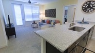 This single-floor residence showcases an open-concept layout that integrates the kitchen and living areas into one cohesive, functional space. The kitchen features a prominent island with a speckled granite countertop, providing ample prep space and a natural gathering point. A built-in sink adds practicality, while the dark cabinetry contrasts elegantly with the light stone surface. Bar-style seating along the island creates a casual dining option that connects the kitchen to the living space.

The living area is generously proportioned, finished with soft wall-to-wall carpeting in a neutral tone that enhances the warm, comfortable atmosphere. Large windows along the far wall allow abundant natural light to fill the space, complemented by a ceiling fan that adds both functionality and a subtle design element. The open sightlines between rooms contribute to an airy, spacious feel throughout.

A hallway visible from the main living area leads to additional rooms, suggesting a well-organized floor plan. The overall aesthetic is clean and contemporary, with soft yellow-toned walls and neutral finishes that create a welcoming, move-in-ready environment.