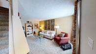 This two-floor residence offers a well-designed interior with distinct spaces that combine comfort and character throughout. The living room serves as a focal point, featuring warm neutral walls accented by a golden yellow feature wall adorned with vintage transit sign artwork. Plush carpeting, a glass-topped coffee table, and ample natural light contribute to a welcoming atmosphere. An open bookcase and layered décor add visual depth to the space.

The kitchen is equipped with white cabinetry, a white range with built-in microwave, and a full-size refrigerator. Light wood-look flooring grounds the space, which transitions seamlessly into the adjacent dining area. The dining room features a round wooden table surrounded by floral-upholstered chairs, a pendant light overhead, and a window that brings in natural light.

The bedroom makes a strong design statement with a dramatic deep-blue wood-paneled accent wall, soft carpeting, and a window allowing natural light to filter in. Two bathrooms complete the home — one featuring a distinctive green marble-style countertop with white cabinetry, and the other offering a clean white vanity with a bright, well-lit mirror. Together, these spaces create a cohesive and characterful home environment.