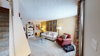 This two-floor residence offers a well-designed interior with distinct spaces that combine comfort and character throughout. The living room serves as a focal point, featuring warm neutral walls accented by a golden yellow feature wall adorned with vintage transit sign artwork. Plush carpeting, a glass-topped coffee table, and ample natural light contribute to a welcoming atmosphere. An open bookcase and layered décor add visual depth to the space.

The kitchen is equipped with white cabinetry, a white range with built-in microwave, and a full-size refrigerator. Light wood-look flooring grounds the space, which transitions seamlessly into the adjacent dining area. The dining room features a round wooden table surrounded by floral-upholstered chairs, a pendant light overhead, and a window that brings in natural light.

The bedroom makes a strong design statement with a dramatic deep-blue wood-paneled accent wall, soft carpeting, and a window allowing natural light to filter in. Two bathrooms complete the home — one featuring a distinctive green marble-style countertop with white cabinetry, and the other offering a clean white vanity with a bright, well-lit mirror. Together, these spaces create a cohesive and characterful home environment.