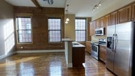 This single-floor loft-style space masterfully combines industrial heritage with contemporary living. Exposed brick walls serve as a defining architectural feature throughout, adding warmth, texture, and a sense of history to the interior. Richly polished hardwood floors run throughout the space, reflecting natural light and enhancing the open, airy atmosphere.

The open-concept kitchen is a standout element, outfitted with warm wood cabinetry, light stone countertops, and a full suite of stainless steel appliances including a range, microwave, and sink. A central kitchen island with a dark base provides additional workspace and serves as a natural gathering point within the layout.

Large, multi-pane black-framed windows line the walls, flooding the space with abundant natural light while offering views of the surrounding brick exterior — a nod to the building's historic mill or warehouse origins. Exposed ductwork and structural timber beams overhead reinforce the loft's industrial character.

Track lighting and pendant fixtures provide well-distributed illumination throughout, complementing the natural light. The overall ambiance is a compelling blend of raw, industrial authenticity and polished modern comfort, making this a distinctive and inviting space.