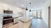 This single-floor residence showcases a thoughtfully designed open-concept layout that seamlessly connects the kitchen and living area. The kitchen is a standout feature, outfitted with crisp white shaker-style cabinetry, granite countertops, a tile backsplash, and a full suite of stainless steel appliances including a range, microwave, dishwasher, and refrigerator. A central island with matching granite provides additional prep and dining space, while two stylish pendant lights hang above, adding a modern touch. Light wood-style flooring runs throughout the main living areas, enhancing the bright, airy feel of the space. Large windows in the living area flood the room with natural light and offer views of the surrounding exterior. A glazed door provides direct access to an outdoor area. The bedrooms are comfortably carpeted and feature windows with blinds, ceiling fans, and generous wall space. The bathroom is clean and contemporary, featuring a soaking tub with tiled surround, a granite-topped vanity with white cabinetry, and light wood-style flooring. Throughout the residence, consistent finishes and neutral tones create a cohesive, modern aesthetic that is both functional and inviting.