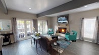 The Veranda Club Room is a thoughtfully designed single-story community space that blends comfort and functionality throughout its open layout. The main lounge area serves as the focal point of the room, anchored by a tile-surround fireplace topped with a wall-mounted television. Warm wood-look flooring runs throughout, complemented by decorative exposed ceiling beams that add architectural character. Vibrant teal accent chairs, a plush charcoal sofa, and a multicolored area rug create a welcoming, contemporary atmosphere. French doors along one wall allow natural light to pour in while providing views of the outdoor area. Adjacent to the lounge, a dedicated dining area features multiple glass-top tables with upholstered seating, accented by a large decorative wall mirror that reflects light and enhances the sense of space. The kitchen is equipped with black appliances, including a range and full-size refrigerator, white cabinetry, and countertops. Pendant lighting illuminates both the kitchen workspace and a bar-height counter with black stools, making it ideal for casual gatherings or catered events. Recessed lighting throughout keeps the space bright and inviting, while neutral gray walls provide a cohesive backdrop for the bold furnishings and décor accents.