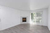 This single-floor space offers a clean, modern aesthetic defined by white walls, light-toned wood-style flooring, and an abundance of natural light throughout. The living area serves as a welcoming focal point, anchored by a sleek wall-mounted fireplace with a white tile surround. A large sliding glass door fitted with vertical blinds opens to an outdoor view of lush greenery, flooding the room with natural light.

The kitchen is thoughtfully designed with a galley-style layout featuring two-toned cabinetry — white upper cabinets paired with sage green lower cabinets — complemented by white countertops and stainless steel appliances including a refrigerator, range, microwave, and dishwasher. Recessed lighting illuminates the space efficiently. A peninsula with bar-height seating potential connects the kitchen to the adjacent dining and entry area, which includes an arched architectural accent and entry door.

The bedroom is carpeted for comfort and includes a walk-in closet with built-in shelving, offering generous storage. A second carpeted room with a baseboard heater provides flexible use as an additional bedroom or office. The bathroom features a white vanity with black hardware, gold-toned faucet, undermount sink, full tub-shower combination, and wood-style tile flooring.