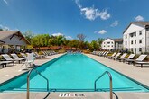 Located at 1905 Riverside Pkwy in Douglasville, Georgia, this thoughtfully designed outdoor amenity area serves a modern multi-building residential community. The centerpiece is a generously sized resort-style swimming pool with crystal-clear turquoise water, surrounded by an expansive concrete pool deck lined with rows of lounge chairs, providing ample space for sunbathing and relaxation. The pool area is framed by wrought iron fencing and offers views of the community's contemporary multi-story residential buildings, which feature white and gray board-and-batten siding with uniformly spaced windows.

Adjacent to the pool, a dedicated outdoor lounge and grilling area is shaded by striking wood-framed pergolas, complete with built-in grilling stations and additional lounge seating — ideal for outdoor entertaining. Shaded patio tables with umbrellas offer quieter spots for dining or gathering.

The grounds are attractively landscaped with manicured lawns, flowering shrubs, ornamental grasses, mulched planting beds, and young trees that add greenery and color throughout. A relaxing hammock area is tucked alongside one of the single-story community buildings, which features a welcoming covered entrance with stone accents and ceiling fans. Mature tree lines along the property perimeter provide a natural backdrop and a sense of privacy.