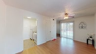 This single-floor unit offers a straightforward and functional layout connecting a living area, kitchen, bathroom, and outdoor space. The living room is the central feature of the unit, finished with light wood-look flooring that extends throughout the main living space. A ceiling fan with an integrated light fixture provides both airflow and ambient lighting, while a textured ceiling adds subtle architectural detail. Natural light enters through a sidelight window flanking the front entry door and a sliding glass door fitted with vertical blinds, which opens to a balcony with outdoor views.

The kitchen is positioned adjacent to the living area and is equipped with white cabinetry, a granite-style laminate countertop, a double stainless steel sink, a freestanding range, and a full-size refrigerator. Tile flooring runs through the kitchen, providing a practical contrast to the living area's wood-look surface.

The bathroom is notably elongated, featuring a granite-style vanity countertop with an undermount sink, white cabinetry below, a large mirror, and a full tub-shower combination with tile surround at the far end. Tile flooring and bright overhead lighting complete the space. Overall, the unit maintains a clean, neutral palette throughout.