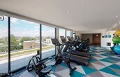 This contemporary fitness center is designed to deliver a premium workout experience within a bright, energizing environment. The space is defined by an expansive wall of floor-to-ceiling windows along one side, flooding the interior with abundant natural light and offering sweeping elevated views of the surrounding cityscape — a motivating backdrop for any workout.

The flooring makes a bold visual statement, featuring a striking geometric pattern in teal, gray, and white that adds a dynamic, modern character to the space. Recessed lighting throughout the clean white ceiling complements the natural light, ensuring the facility remains well-lit at all times.

The cardio zone is generously appointed with multiple treadmills, elliptical trainers, stationary upright and recumbent bikes, a rowing machine, and a Peloton-style cycle bike. A dedicated strength and functional training area houses a Matrix cable machine system, an adjustable weight bench, and a well-stocked rack of medicine balls, resistance bands, and exercise mats in various sizes.

A separate alcove provides additional space for focused training and features a sliding glass door opening to an outdoor terrace, further enhancing the sense of openness. A water station is conveniently accessible within the space. The overall ambiance is clean, modern, and energizing, making this fitness center a standout amenity.