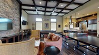 Located at 100 Village Way in Goleta, California, this single-story community clubhouse is designed to serve as a central social hub. The space is unified by rich, dark hardwood-style flooring and a striking coffered ceiling with bold dark wood beams, lending the interior a warm yet sophisticated character throughout.

The main gathering area functions as a game and lounge room, anchored by a full-size pool table beneath a linear pendant light fixture. A ping-pong table provides additional recreation, while comfortable seating—including a leather sofa and upholstered accent chairs—creates an inviting lounge arrangement. A dramatic stacked-stone accent wall serves as a focal point, housing a wall-mounted flat-screen television. Clerestory windows allow natural light to filter in while maintaining privacy.

A dedicated bar-height seating nook with round tables and stools offers a secondary gathering spot, complete with its own wall-mounted television—ideal for watching games or socializing in smaller groups.

The full kitchen is well-appointed with dark wood cabinetry, white quartz countertops, stainless steel appliances including a professional-grade range with an overhead vent hood, a large refrigerator, and a center island with a gas cooktop and teal leather bar stools. A mosaic tile backsplash and stone accent wall add artisanal detail, completing this highly functional and stylish communal space.