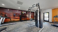 Located at 1333 N Dysart Rd in Avondale, Arizona, this fitness center offers a well-equipped exercise space designed to support a complete workout routine. The room is finished with durable carpet tile flooring and features a bold, energizing color scheme — vibrant orange accent walls contrast with clean white surfaces, creating a motivating environment for exercise.

The space is outfitted with a variety of strength and cardio equipment, including a multi-function cable machine, a Smith machine with an adjustable bench, treadmills, and a stationary spin bike. A dedicated training zone along one wall features a tiered dumbbell rack and additional free weights, keeping equipment organized and accessible. A large, dramatic motivational mural with the phrase "Be Fearless" anchors the space visually, reinforcing the fitness-focused atmosphere.

Recessed lighting throughout provides consistent illumination, while a window along one wall allows natural light to filter in, offering views of the surrounding outdoor greenery. A wall-mounted television and informational signage add functional touches. Double glass doors at the entrance provide easy access and contribute to an open, welcoming feel. The layout is thoughtfully arranged to allow users to move freely between equipment, making the most of the available floor area.