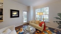 Located at 1316 Town Center Dr in Pflugerville, Texas, this single-story residence offers a thoughtfully designed layout with consistent, quality finishes throughout. The living area greets visitors with rich hardwood-style flooring, elegant crown molding, and generous natural light streaming through large windows with blinds that frame views of the surrounding greenery. The space accommodates comfortable seating and decorative accents in a bright, welcoming atmosphere.

The dining area makes a striking statement with a bold navy accent wall, track lighting overhead, and crown molding that ties the room together. The kitchen is outfitted with dark espresso cabinetry, granite-style countertops, black appliances including a range, microwave, and refrigerator, a tile backsplash, and light tile flooring. A window above the sink allows natural light to fill the workspace.

The bedroom features plush carpeting, crown molding, a ceiling fan, and a large arched window that brings in ample daylight. An additional carpeted room with a window offers flexibility as a study or secondary sitting space. The bathroom is finished with dark cabinetry, granite-topped vanity, tile flooring, and a fully tiled tub-shower surround, completing this well-appointed home.